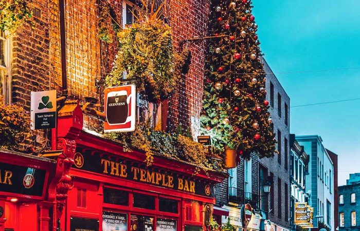 Brightly lit, festive exterior of The Temple Bar pub decorated with glowing curtain lights across the brick facade, lush greenery, hanging shamrock and Guinness signs, and a tall Christmas tree adorned with red and gold ornaments under a dusk twilight sky.