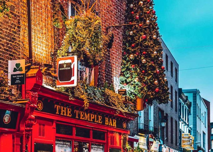 Brightly lit, festive exterior of The Temple Bar pub decorated with glowing curtain lights across the brick facade, lush greenery, hanging shamrock and Guinness signs, and a tall Christmas tree adorned with red and gold ornaments under a dusk twilight sky.
